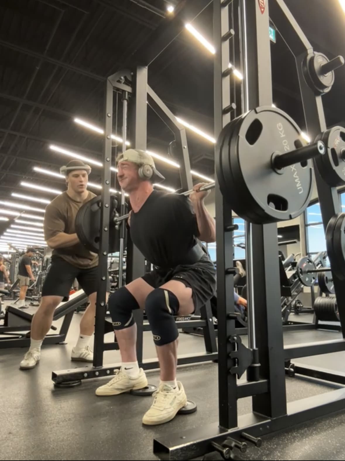 Man performing a squat on a Smith machine with heels elevated and a spotter nearby.